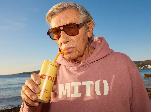 Candid lifestyle product photograph. An older silver-haired man with a weathered, characterful face wearing large tinted sunglasses, drinking from a straw inserted into a yellow MIT() soda can held in his hand. He is wearing a dusty rose pink MIT() hoodie with the cream "MIT()" logo clearly visible on the chest. Medium close-up shot, slightly low angle. Sunny coastal outdoor setting — blue sky and water in the background, warm golden natural light. Relaxed, confident, effortlessly cool expression. The yellow MIT() can with red "MIT()" lettering and "SPARKLING ENERGY ZERO SUGAR" text is clearly visible. Candid editorial lifestyle photography style. No extra text or graphics.
Do not reproduce incidental presentation headers, view names, palette labels, captions, annotations, borders, grids, or layout from the reference images. Preserve writing from a reference only when this prompt explicitly requires that exact in-world text or graphic.